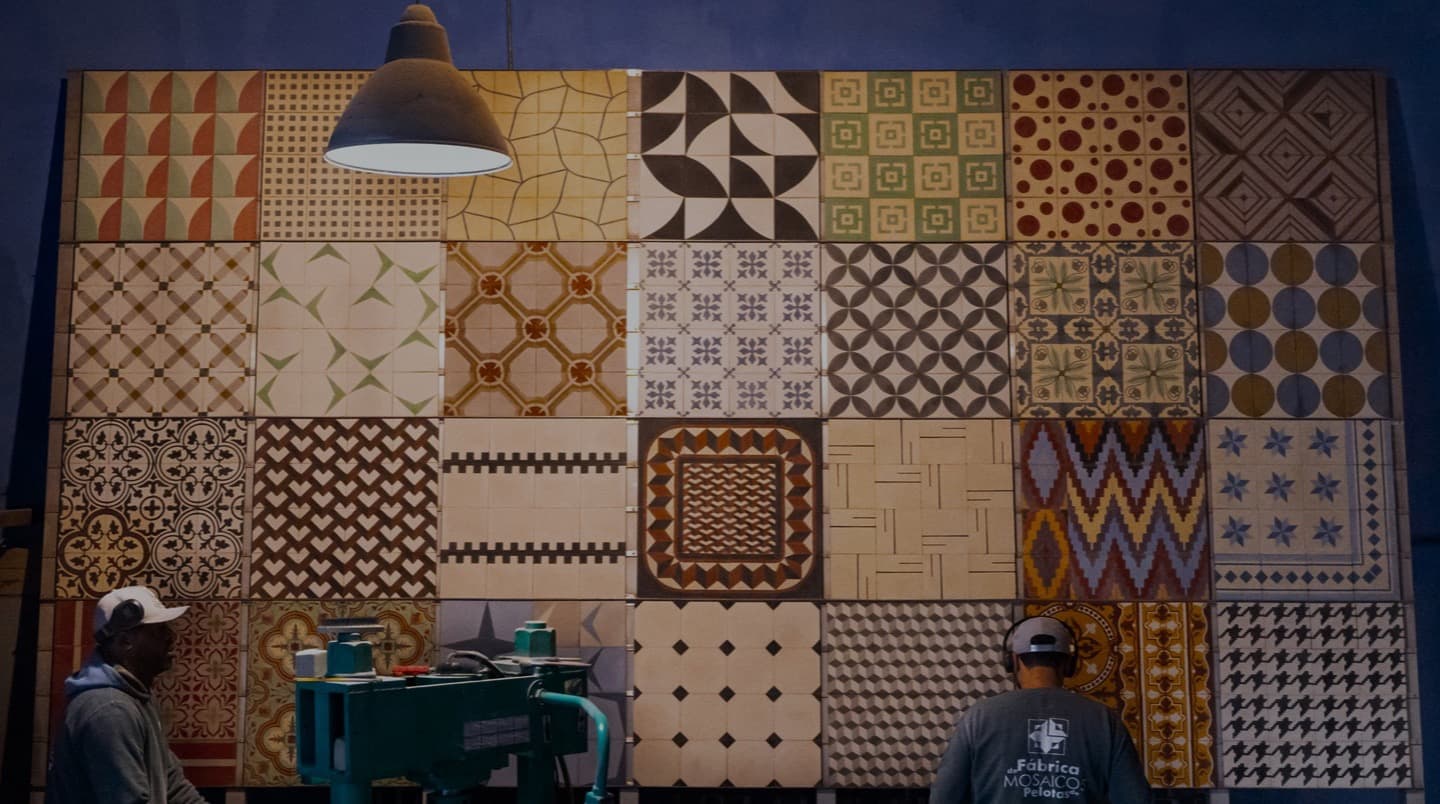 Painel do acervo com diversos padrões de ladrilhos hidráulicos e dois artesãos
